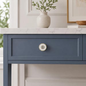 May include: A round cabinet knob featuring a white center with a gold-toned beaded rim and a central clear crystal accent. The knob is mounted on a blue drawer front with a white marble countertop surface visible above the drawer unit.