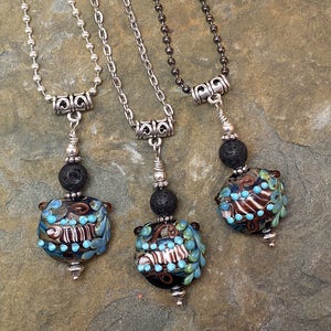 May include: Three silver chain necklaces with black lava stone beads and colorful glass pendants. The pendants feature a blue, green, and white floral design.