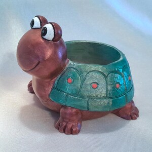 May include: A copper-colored turtle planter with a green shell and red dots. The turtle has large, white eyes and a smiling mouth. The planter is perfect for small plants or succulents.