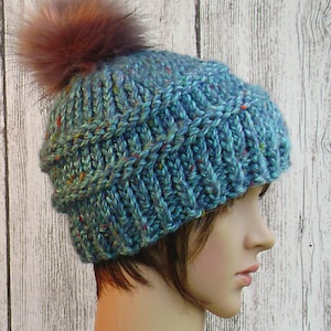 May include: A teal knit hat with a ribbed design and a brown faux fur pom-pom. The hat has flecks of red, yellow, and blue throughout the yarn. The hat is shown on a mannequin head.