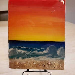 May include: A square resin art piece depicting a sunset over the ocean. The top half of the piece is a gradient of red, orange, and yellow, with a few small white birds flying in the sky. The bottom half of the piece is a blue ocean with white waves crashing on a sandy beach. There are small seashells and a starfish on the beach.