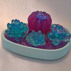 May include: A light blue ceramic dish with a purple and blue resin succulent arrangement. The arrangement features a large purple pumpkin-shaped succulent and three smaller blue succulents.