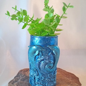 May include: A blue ceramic vase with a dolphin design and a swirling wave pattern. The vase is filled with green leafy plants.