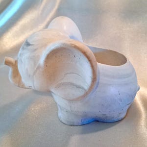 May include: A ceramic elephant planter in shades of white, gray, and beige. The elephant has large ears and a trunk, with a hollowed-out back for planting small plants or succulents. The planter has a marbled effect.