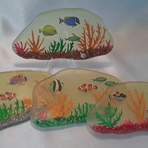 May include: Four resin coasters with a beach theme. Each coaster features a different design of colorful fish and coral reefs on a light yellow background.