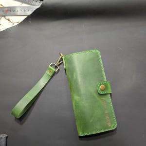 Könnte beinhalten: Eine grüne Ledergeldbörse mit passender Handschlaufe, einem Messing-Druckknopfverschluss und einem eingeprägten Namen auf der Vorderseite. Ein Metalllineal ist oben sichtbar und zeigt Maße in Zentimetern, wobei die 20-cm-Markierung deutlich zu sehen ist.