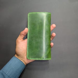 Könnte beinhalten: Ein rechteckiges Portemonnaie aus glattem, waldgrünem Leder mit sichtbaren, farblich abgestimmten Nähten entlang der Kanten. Das Portemonnaie hat ein schlichtes Design mit matter Oberfläche und abgerundeten Ecken in einem einheitlichen Grünton.