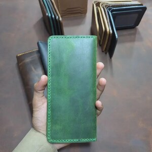 May include: A rectangular green leather wallet featuring visible stitched edges. The wallet has a smooth, matte finish with subtle colour variations across the surface. Several other leather wallets in brown and black tones are visible in the background.