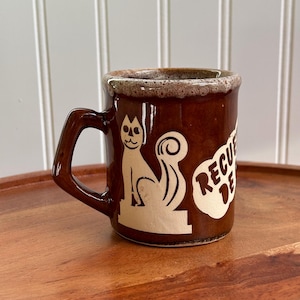 May include: A brown ceramic mug with a speckled rim and a handle. The mug features a cream-colored graphic of a stylized cat sitting on a base, positioned next to a cream-colored shape containing the partial text RECU DE.
