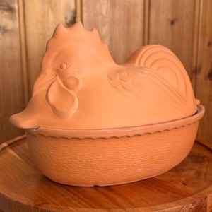 May include: A terracotta chicken-shaped lidded casserole dish with a textured base and a molded rooster lid. The piece features a matte finish in a warm orange-brown clay tone, with detailed carvings of feathers and facial features on the lid.