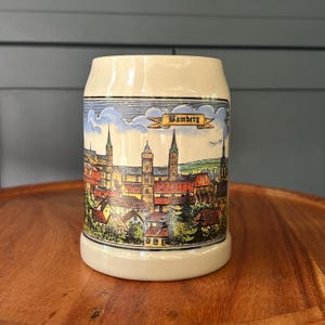 May include: A cylindrical ceramic beer stein with a cream-colored base. The exterior features a colorful, detailed illustration of the city of Bamberg, Germany, including various buildings, church spires, and trees, with the text Bamberg in a banner.
