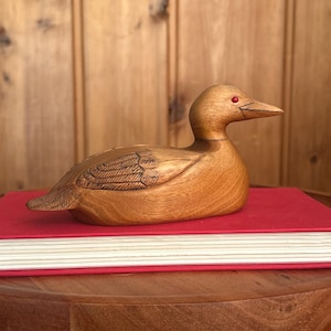 May include: A hand-carved wooden duck figurine featuring a smooth light brown finish, detailed etched wing patterns, and a single small red bead eye. The duck is positioned in a resting pose with its head turned slightly to the right side.