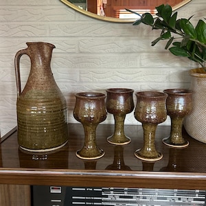 May include: A ceramic pitcher and four matching stemmed goblets featuring a two-tone glaze with a dark brown speckled upper section and a muted olive green lower section. The pitcher has a curved handle and a flared spout, while the goblets have wide bowls.
