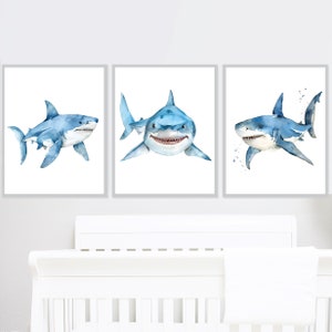 May include: Set of three watercolor paintings featuring blue sharks on a white background. The sharks are depicted in different poses, with one swimming towards the viewer, one swimming away, and one swimming sideways.