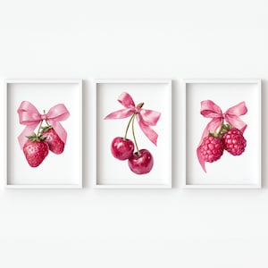 Op de afbeelding: Set van drie ingelijste aquarelprints met rode vruchten: aardbeien, kersen en frambozen. Elke vrucht is versierd met een delicate roze strik. De kunstwerken staan tegen een witte achtergrond, wat een frisse en elegante uitstraling creëert. Afmetingen in centimeters.