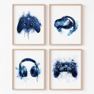May include: Four framed watercolor prints featuring gaming accessories. The prints depict a blue game controller, VR headset, headphones, and another game controller, all against a white background with blue paint splatter effects. The frames are light brown.
