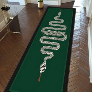 Puede incluir: Una alfombra rectangular de pasillo con fondo verde oscuro y borde negro. Un gráfico de serpiente larga y sinuosa con un patrón geométrico de triángulos blancos y negros recorre el centro, con una lengua roja bífida en cada extremo.