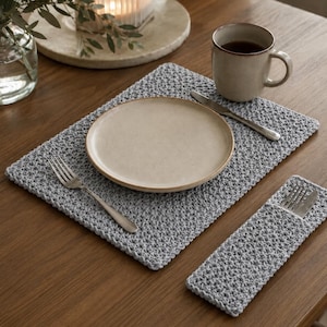 May include: A rectangular light gray crocheted placemat and a matching rectangular cutlery holder. The placemat features a textured stitch pattern and holds a round beige ceramic plate, a silver fork, and a silver knife. A matching gray crocheted sleeve contains a second silver fork and knife.