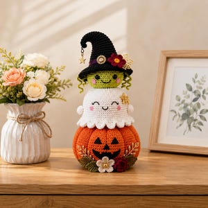 May include: A stacked crochet figurine featuring a bright orange jack-o'-lantern base, a white ghost in the middle, and a green-faced witch wearing a black pointed hat with floral accents and a small gold star charm hanging from the brim.