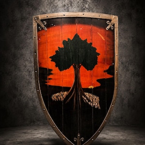 May include: A wooden heater shield featuring a painted scene of a black tree silhouette against a vibrant orange and red sunset background. The shield has a dark wooden frame reinforced with metal trim and rivets along the edges and at the base.