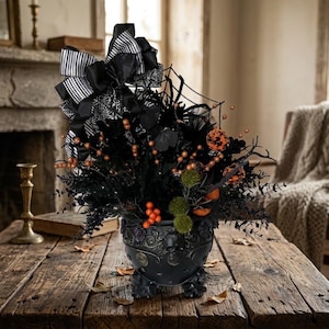 May include: A black cauldron-shaped planter featuring embossed skull and floral details, filled with black foliage, orange berry clusters, green spherical accents, and a large black and white striped bow with a spiderweb pattern on top.