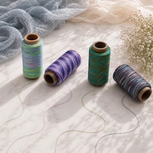 May include: Four cylindrical spools of metallic thread in varying color combinations: one light blue and green, one purple and blue, one teal and gold, and one dark blue and red. Each spool has a brown cardboard core and a single loose thread tail.