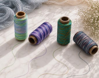 Könnte beinhalten: Vier zylindrische Garnrollen aus Metallic-Garn in verschiedenen Farbkombinationen: hellblau und grün, lila und blau, petrol und gold sowie dunkelblau und rot. Jede Spule hat einen braunen Pappkern und einen einzelnen losen Faden.