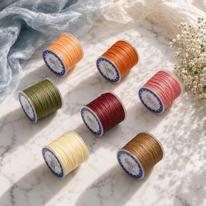 May include: Six spools of thread in various colors including light orange, dark orange, olive green, deep red, pink, cream, and brown. Each spool features a white circular label with blue text and a small graphic icon on the side of the plastic core.
