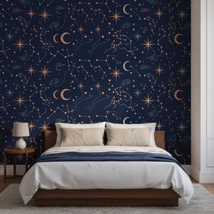 May include: Wallpaper featuring a dark navy blue background with a repeating pattern of light peach and white constellations, stars, crescent moons, and small swirling clouds. The design is displayed on a wall behind a bed with beige pillows.