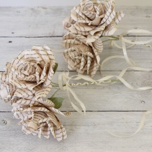 Puede incluir: Dos rosas de papel hechas con p&aacute;ginas de libros con tallos de cinta de color crema. Las rosas est&aacute;n sobre un fondo de madera blanqueada.