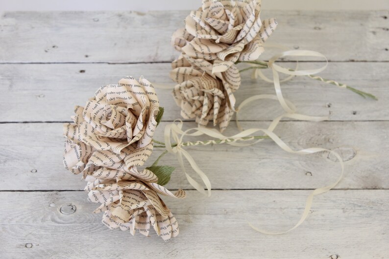 Puede incluir: Dos rosas de papel hechas con p&aacute;ginas de libros con tallos de cinta de color crema. Las rosas est&aacute;n sobre un fondo de madera blanqueada.