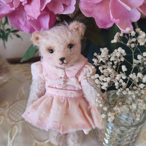 May include: A small, cream-colored teddy bear wearing a pink dress with white polka dots and a pearl button. The bear is sitting on a white lace tablecloth with pink flowers and white baby's breath in the background.