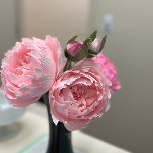May include: A bouquet of three pink paper peonies in a black vase. The peonies are made of crinkled paper and have a realistic appearance.