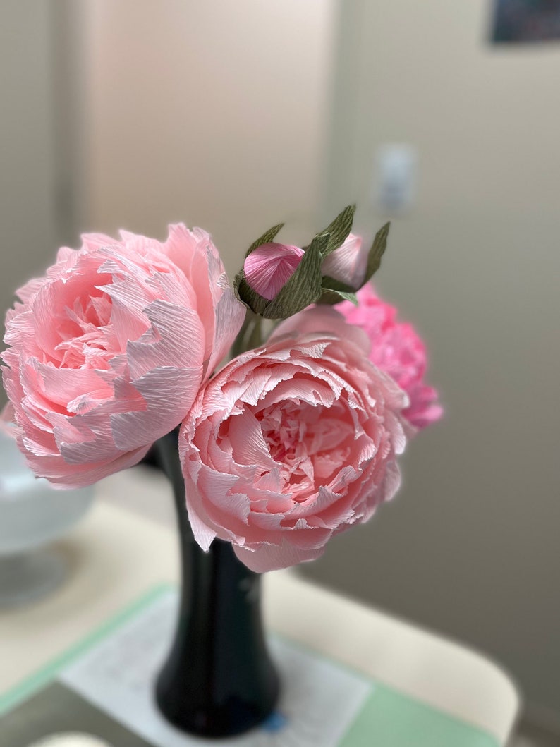 May include: A bouquet of three pink paper peonies in a black vase. The peonies are made of crinkled paper and have a realistic appearance.
