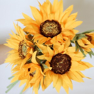 May include: A bouquet of six paper sunflowers in various stages of bloom. The sunflowers are a bright yellow color with brown centers. The bouquet is arranged in a hand-tied style.