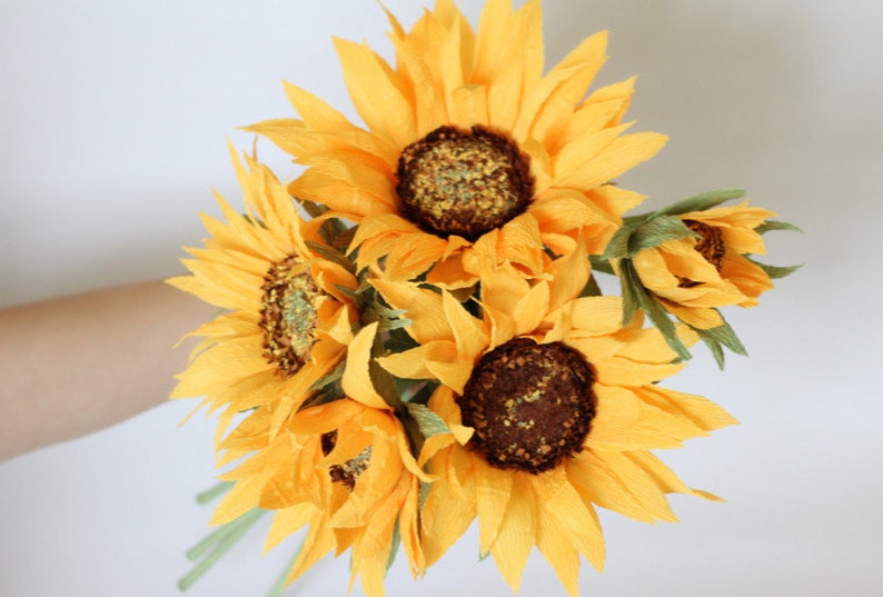 May include: A bouquet of six paper sunflowers in various stages of bloom. The sunflowers are a bright yellow color with brown centers. The bouquet is arranged in a hand-tied style.