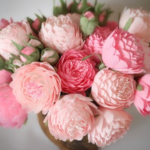 May include: A bouquet of pink paper peonies in a brown vase. The peonies are arranged in a circular pattern and have a delicate, ruffled texture.