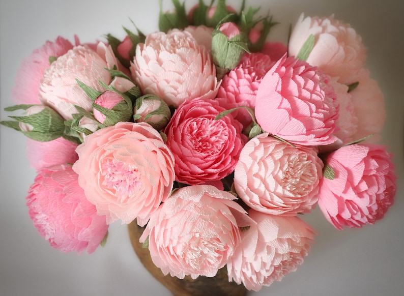 May include: A bouquet of pink paper peonies in a brown vase. The peonies are arranged in a circular pattern and have a delicate, ruffled texture.
