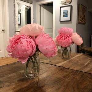 May include: Two glass vases filled with pink paper peonies. The vases are on a wooden table.