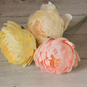 May include: Three paper flowers in shades of yellow and pink. The flowers are arranged in a bouquet and are sitting on a wooden surface.