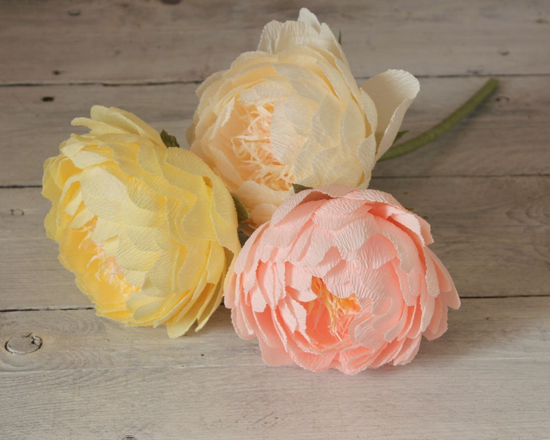 May include: Three paper flowers in shades of yellow and pink. The flowers are arranged in a bouquet and are sitting on a wooden surface.