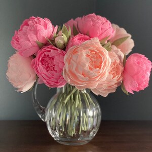 May include: A bouquet of pink and peach paper peonies arranged in a clear glass pitcher with a textured surface.