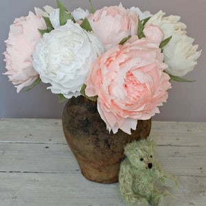 May include: A bouquet of pink and white paper peonies in a rustic brown ceramic vase. The vase is sitting on a white wooden surface.