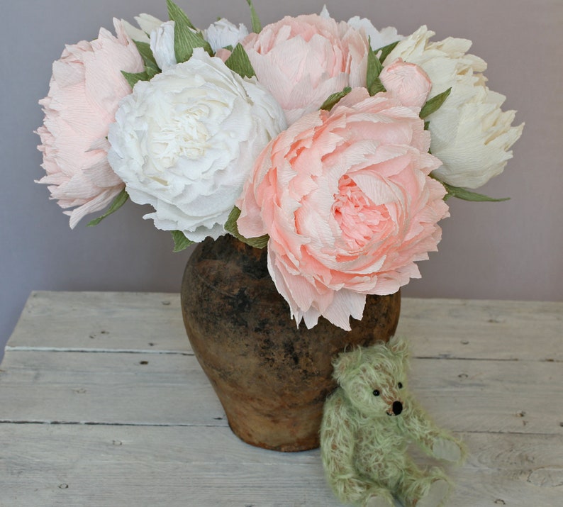 May include: A bouquet of pink and white paper peonies in a rustic brown ceramic vase. The vase is sitting on a white wooden surface.
