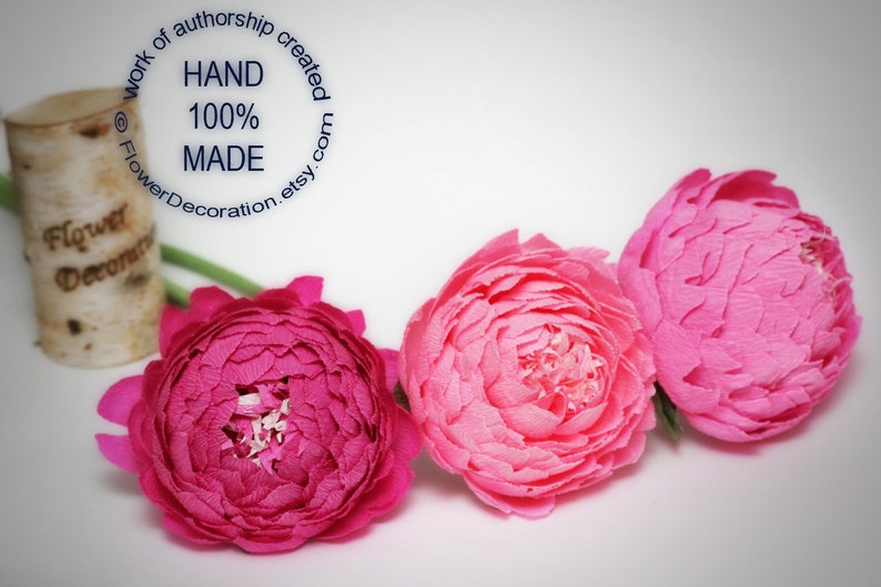 May include: Three paper flowers, one pink and two magenta, arranged in a row on a white surface. The flowers are handmade and have a delicate, layered appearance.