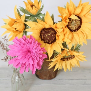 May include: A bouquet of paper flowers in a brown vase. The bouquet includes several yellow sunflowers and one pink flower.