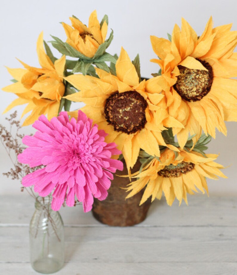 May include: A bouquet of paper flowers in a brown vase. The bouquet includes several yellow sunflowers and one pink flower.