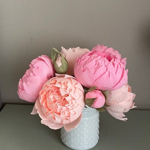 May include: A bouquet of pink and peach paper peonies in a white ceramic vase with a textured surface.