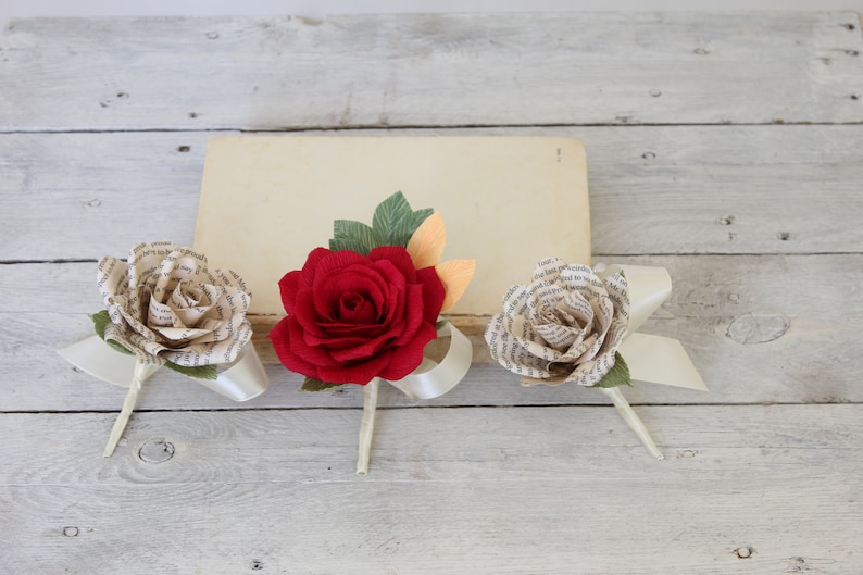 Puede incluir: Tres rosas de papel, una roja y dos de color crema, con detalles de cinta blanca. Las rosas est&aacute;n hechas de p&aacute;ginas de libros.