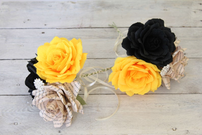 Puede incluir: Dos rosas de papel amarillas y una rosa de papel negra con una rosa de p&aacute;gina de libro en el centro. Las rosas est&aacute;n sobre un fondo de madera blanco.